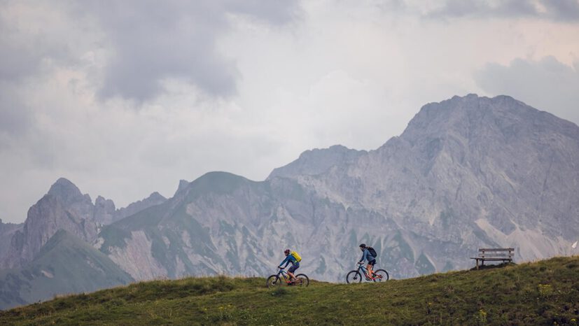 Two mountain bikers ride across a green meadow with the mountain scenery of Faschina in the background.