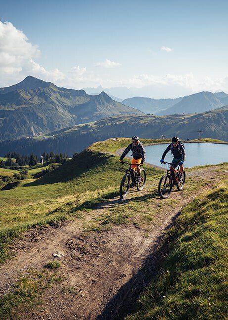 Two mountain bikers in Damüls/Faschina ride up a forest path. In the background there is a lake and the mountain backdrop.