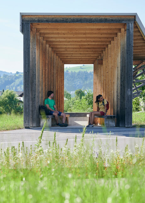 Two hikers are sitting at a bus stop in the middle of nature, waiting for the bus.