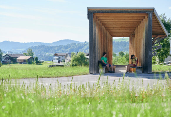 Two hikers are sitting at a bus stop in the middle of nature, waiting for the bus.